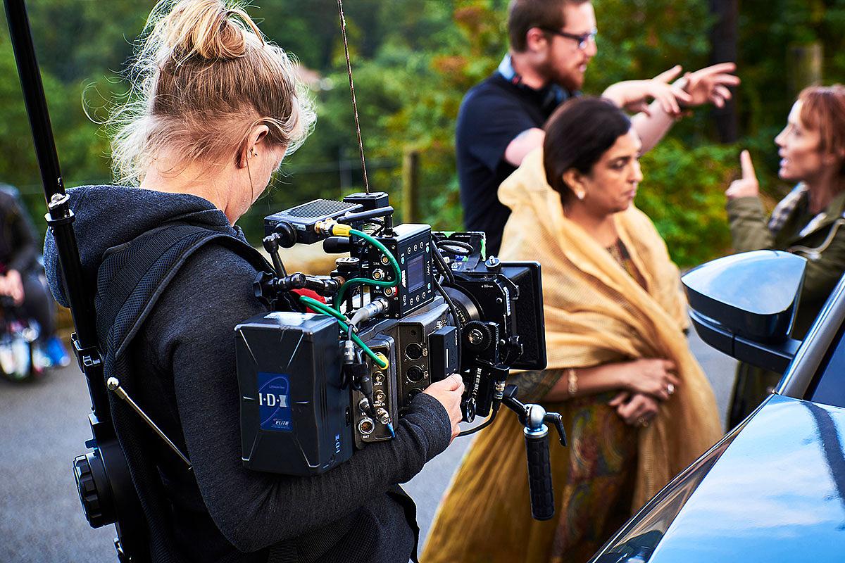 A camera operator is pictured from behind, holding a camera towards two people that are being directed for a shot.