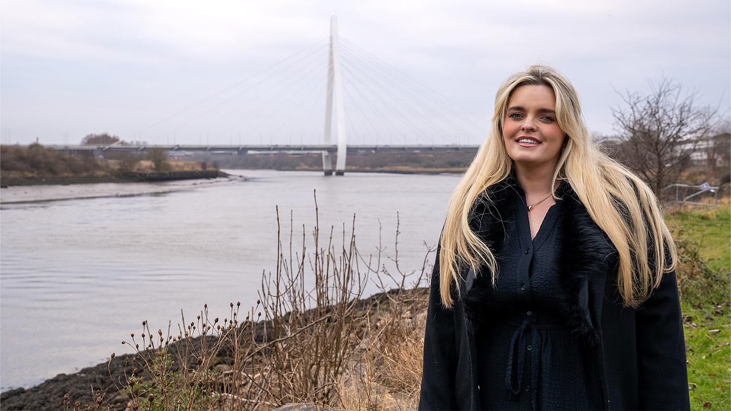 Photograph of Carley Armstrong standing in front of the Northern Spire Bridge in Sunderland on an overcast day. Carley has long, blonde hair, wears a black coat and is smiling.