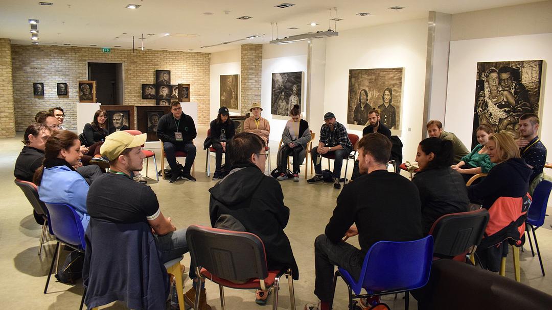 Photograph of a filmmaker roundtable discussion at the Bolton Film Festival, in a gallery space.  A circle of individuals are seated in chairs facing inwards, engaged in conversation.