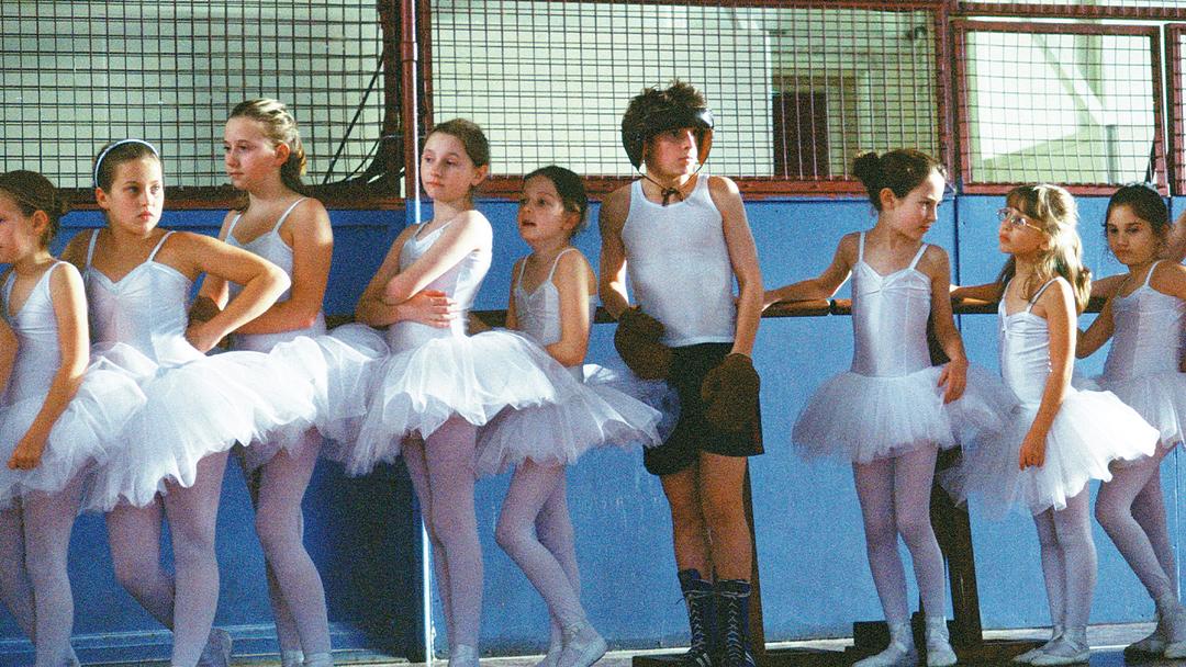 Still from Billy Elliot shows line of young girls dressed in ballerina costumes with young boy at centre wearing boxing helmet