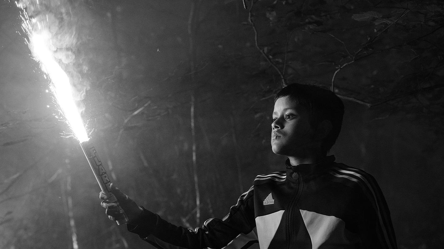 A young man holds a lit flare in a dark wooded area