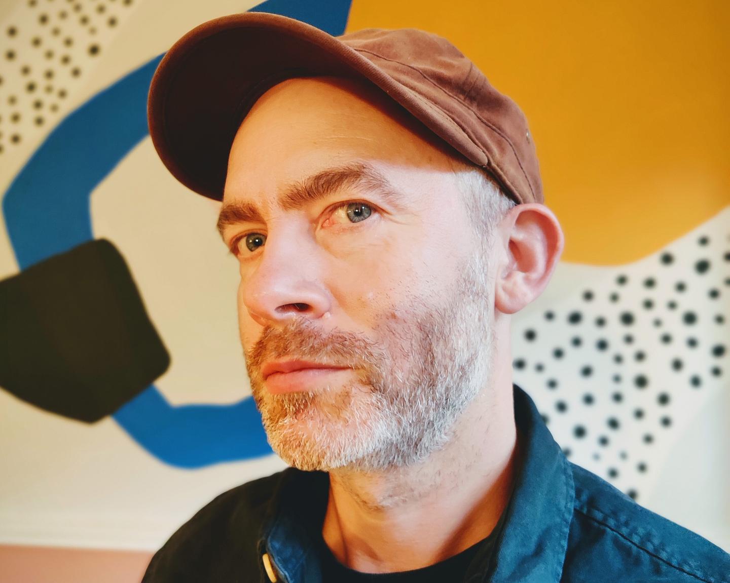 Headshot of Sean Mckenna, a man with light grey facial hair, wearing a brown baseball cap.