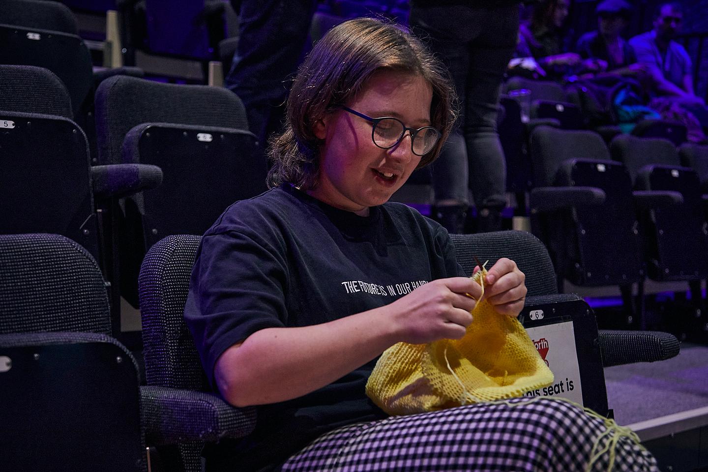 A person with glasses knits while sitting in a cinema seat.