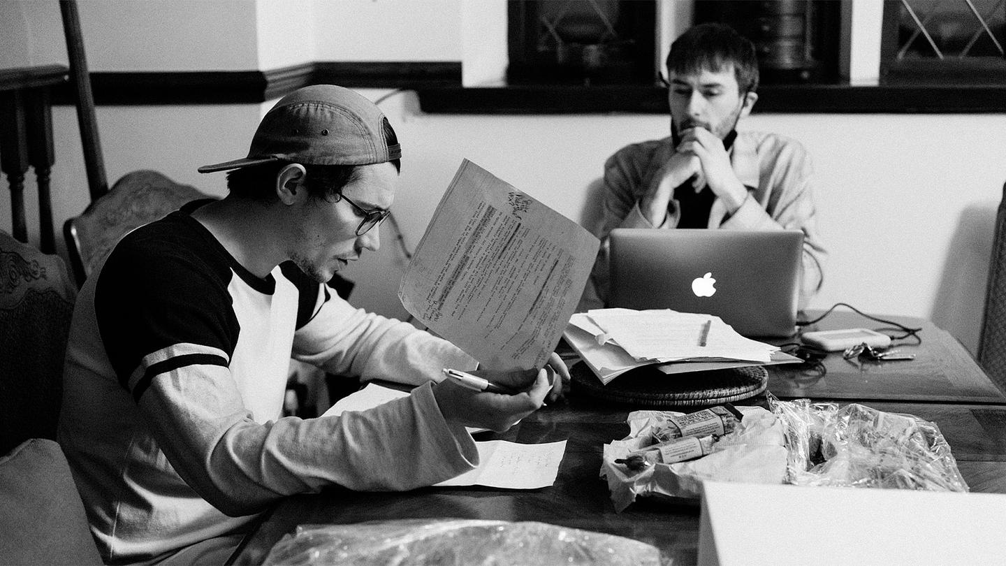 Black and white photograph of two men – Joseph Simmons and Lewis Coates – work intently at a table covered in papers. Joe (wearing a baseball cap and glasses) makes edits to a sheet of paper, whilst Lewis works on a laptop.