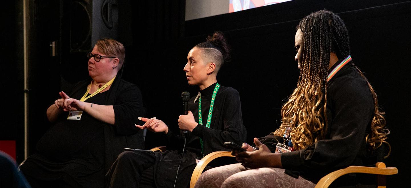 Three people sit on stage taking part in a panel discussion. One is holding a microphone and the other is doing British Sign Language.