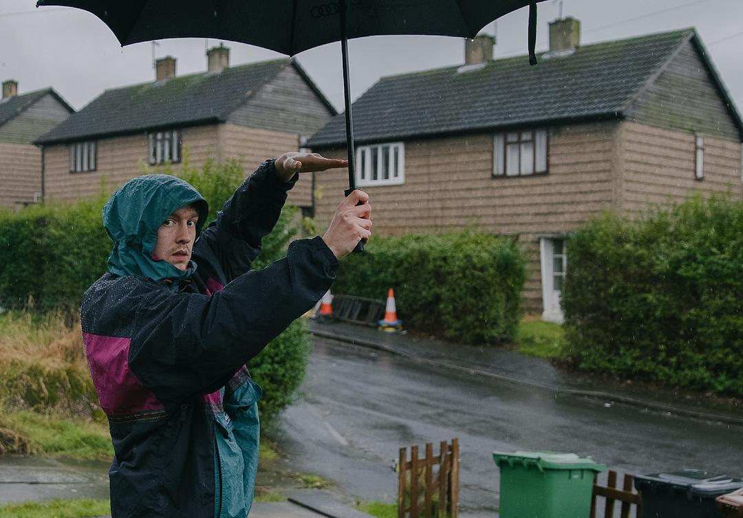 Alfie Barker is wearing a blue and maroon, hooded-raincoat. He is holding an umbrella above his head and using one hand to measure against the pole of the umbrella.