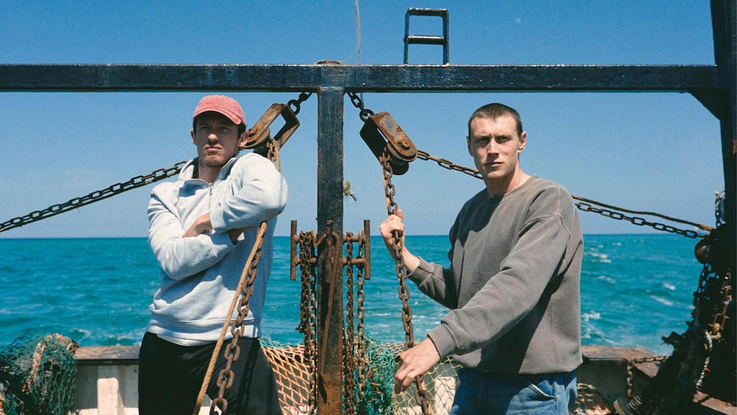 Still from Rose of Nevada shows two men standing on a working boat amidst rusty fishing gear, with a blue sea and clear sky behind.