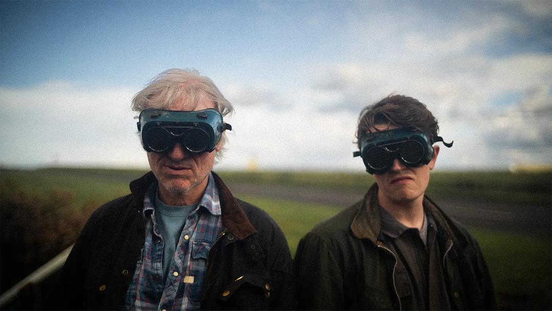 Photograph still from Giants features two men standing side by side in a field, wearing dark goggles that cover their eyes.