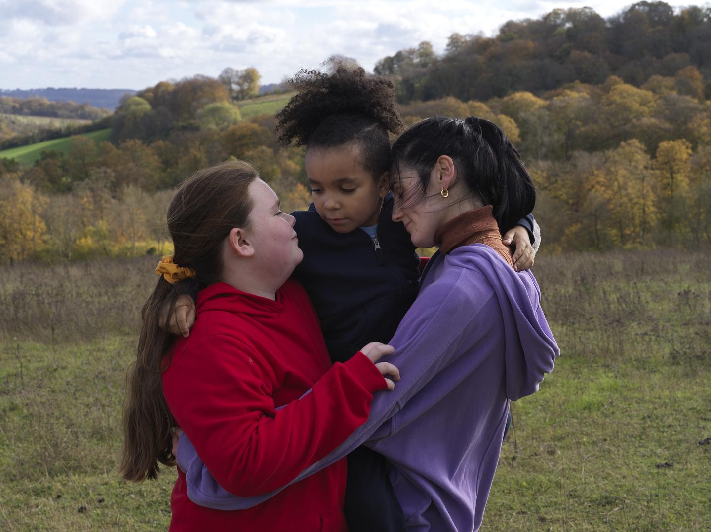 Photograph of three young girls in a tight embrace, in an outdoor field.