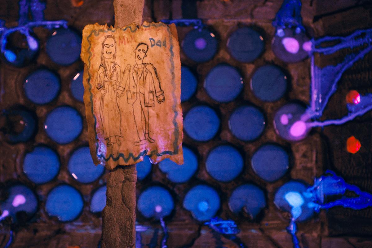 A child's drawing of their parents is held above a vat of glowing blue liquid in a scene from FUTURE PERFECT