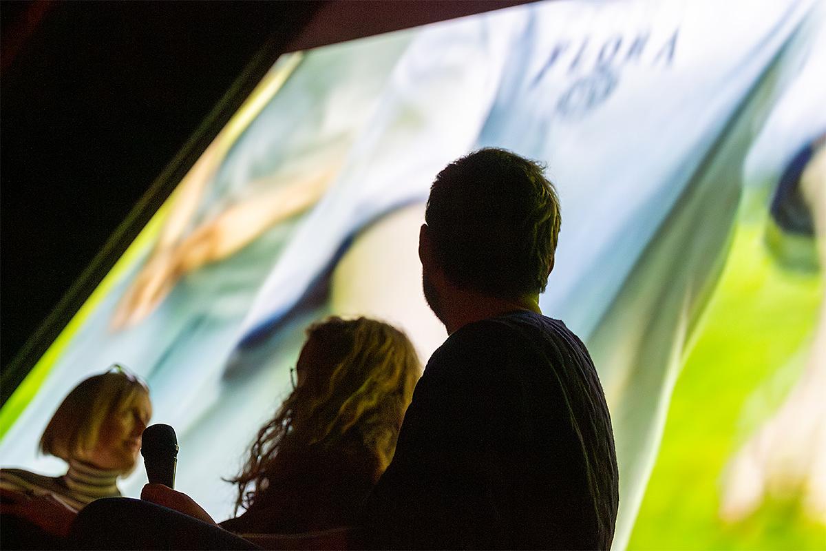 3 people their back to the camera are looking at cinema screen. On the screen there are two people in a field, wearing football shirts