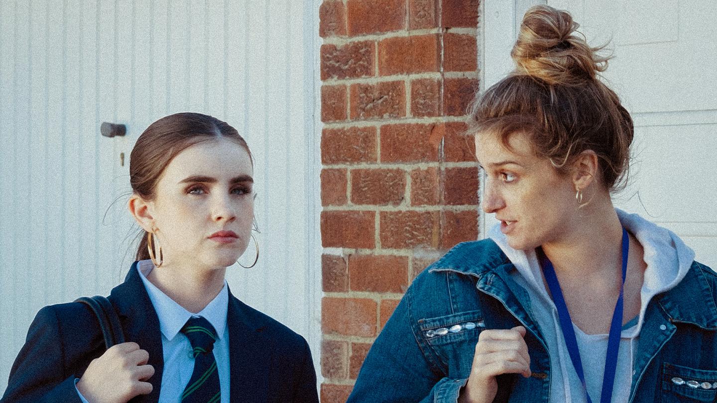 Still from Nowt features a young girl (wearing a school uniform) and a woman standing in front of a brick wall and white door.