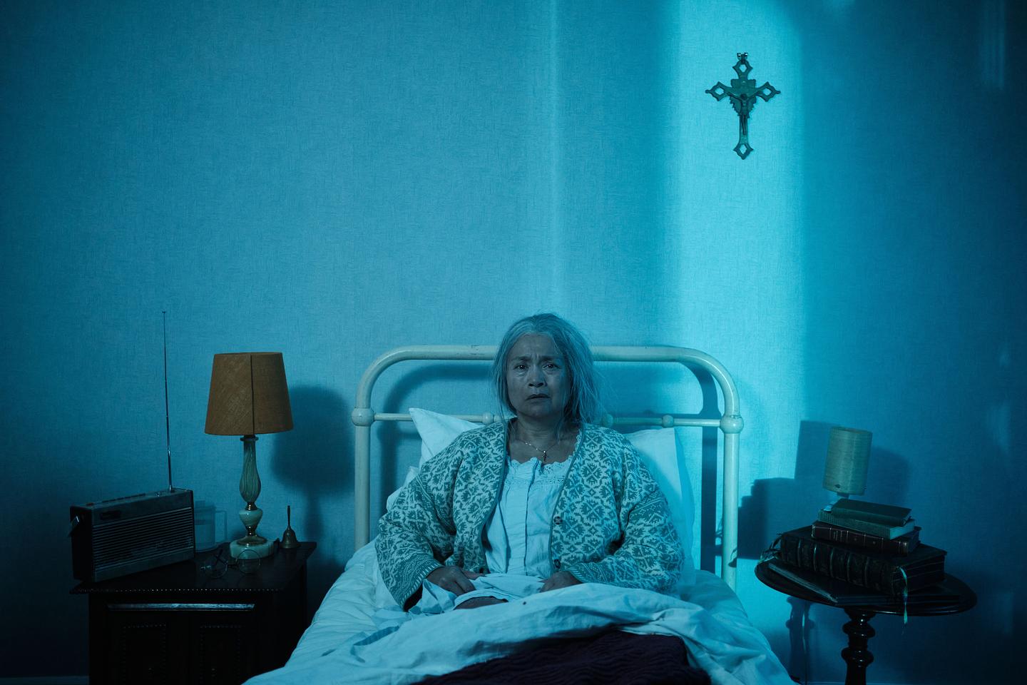 Photo still from Habituation: an older woman sits down on a bed looking sadly into the camera