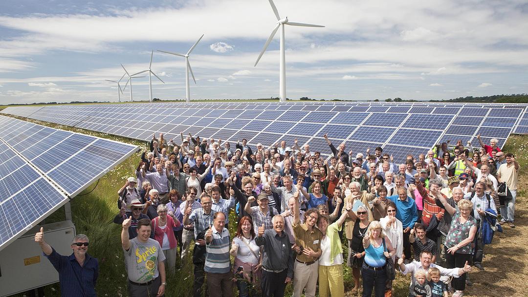 A large group of people cheer and give thumbs-up, posing in front of solar panels and wind turbines.