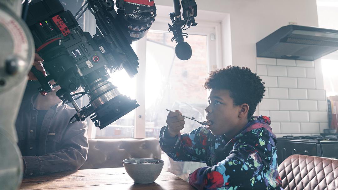 Photograph still from Softy shows a young black boy sitting at a kitchen table, eating cereal. He is being filmed by a professional film crew; a large camera and other filming equipment are visible above him.