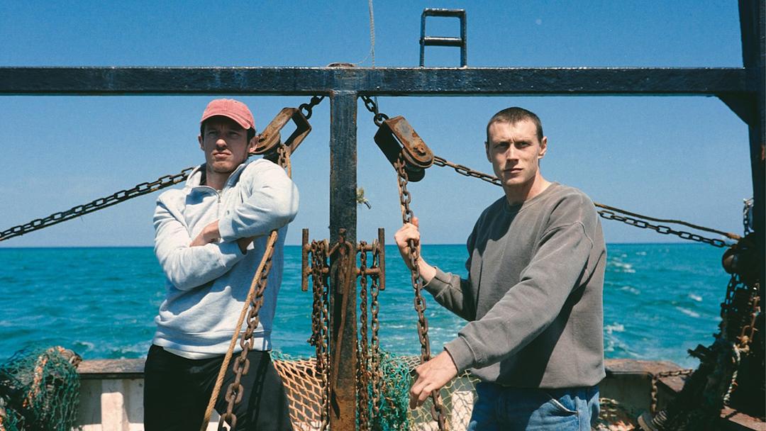 Still from Rose of Nevada shows two men standing on a working boat amidst rusty fishing gear, with a blue sea and clear sky behind.