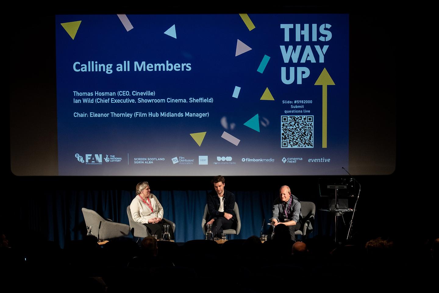 Three panel speakers sit in armchairs on a cinema theatre stage. Behind them, a slide on screen reads: This Way Up, Calling all Members