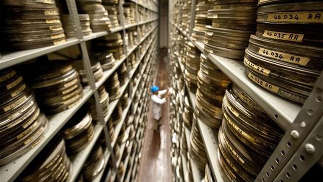 Photograph of BFI Archive: a person wearing a blue hard hat works in a long aisle filled with shelves stacked high with film canisters.