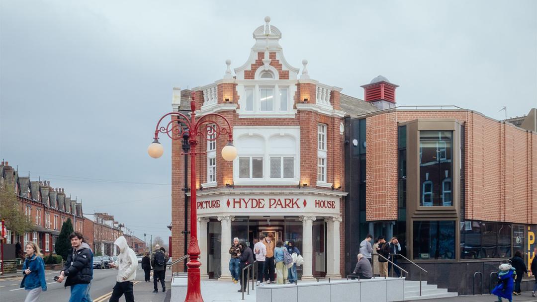 Exterior photograph of Hyde Park Picture House, a red brick building with white columns and window frames, with the cinema’s name displayed in red lettering. A group of people are gathered near the entrance.