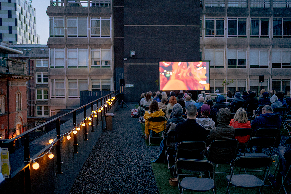 A group of people sat on a rooftop watching a film on a large screen