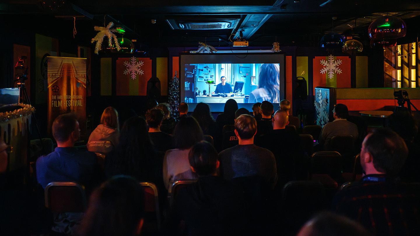 Photograph from The Bay International Film Festival horror screening features a film screening in a small dimly lit venue: a group of people are seated, facing a screen that displays a scene from a movie featuring a man sitting at a desk in an office.