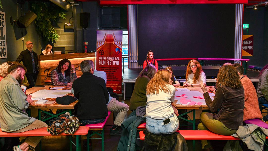 Session from The Bay Film Festival: a group of people are seated around long, communal tables, engaging in discussion.