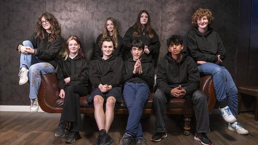 Photograph of a group of eight young adults, either posing against a wall or sitting on a leather sofa.